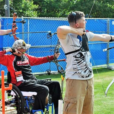 Man standing shooting archery next to man seated in wheelchair shooting archery