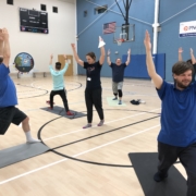 Six people in a circle standing on yoga mats at Bridge 2 Sports Goalball practice on February 10. Each person is standing with one leg outstretched to the back and both arms outstretched above their heads. Jessica Long is in the center of the circle providing direction.