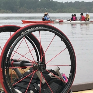 4 people in kayaks in the distance on the lake. In the foreground, is a wheelchair on the dock