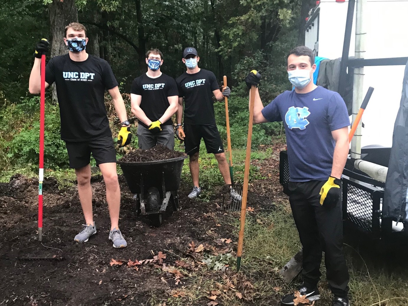 4 male college students standing in mulch holding shovels, rakes and a wheelbarrow. All are wearing masks.