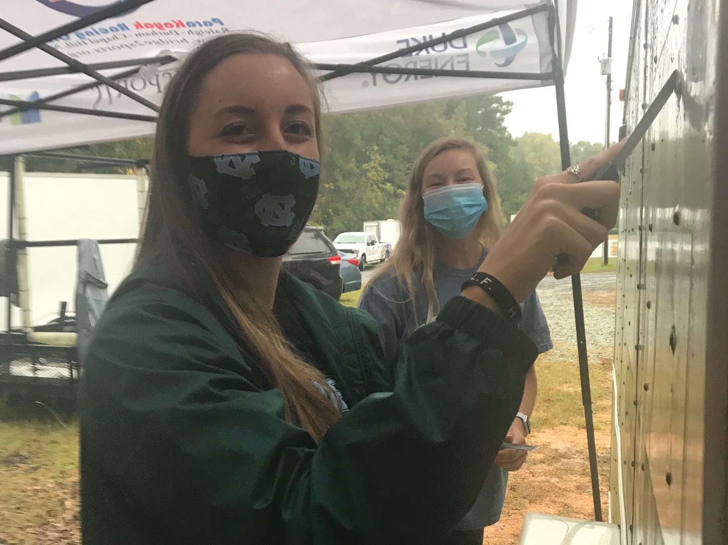 2 female college students using scrapers to remove decals off Bridge 2 Sports trailer. Both are wearing masks and looking at the camera.