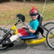 Boy sidts on yellow handcycle smiling at camera. He has a below the knee left leg amputation. He is wearing sunglasses and a helmet with mohawk spikes on it.