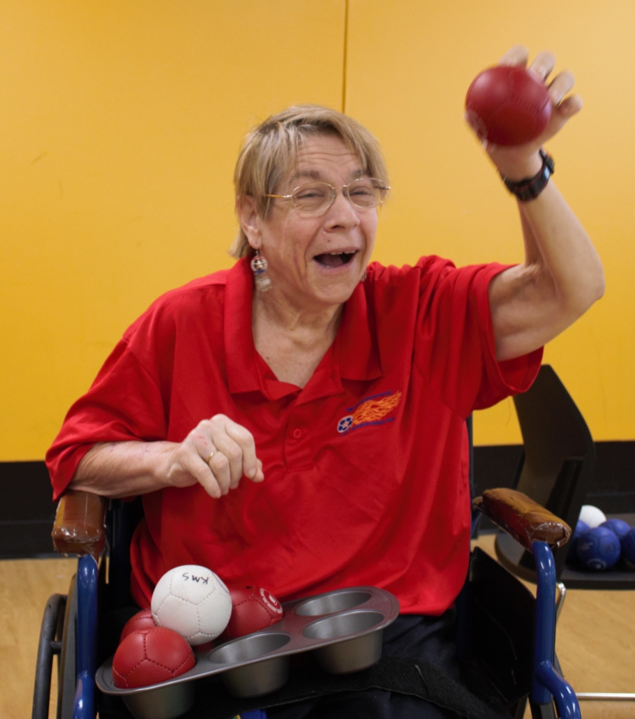 Karen Stallings sits in wheelchair smiling with raised arm throwing a boccia ball