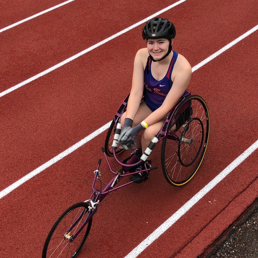 High school student, Jillian, sits in racing chair on track smiling wearing helmet and team tanktop uniform