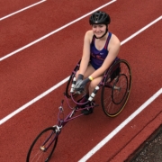 High school student, Jillian, sits in racing chair on track smiling wearing helmet and team tanktop uniform