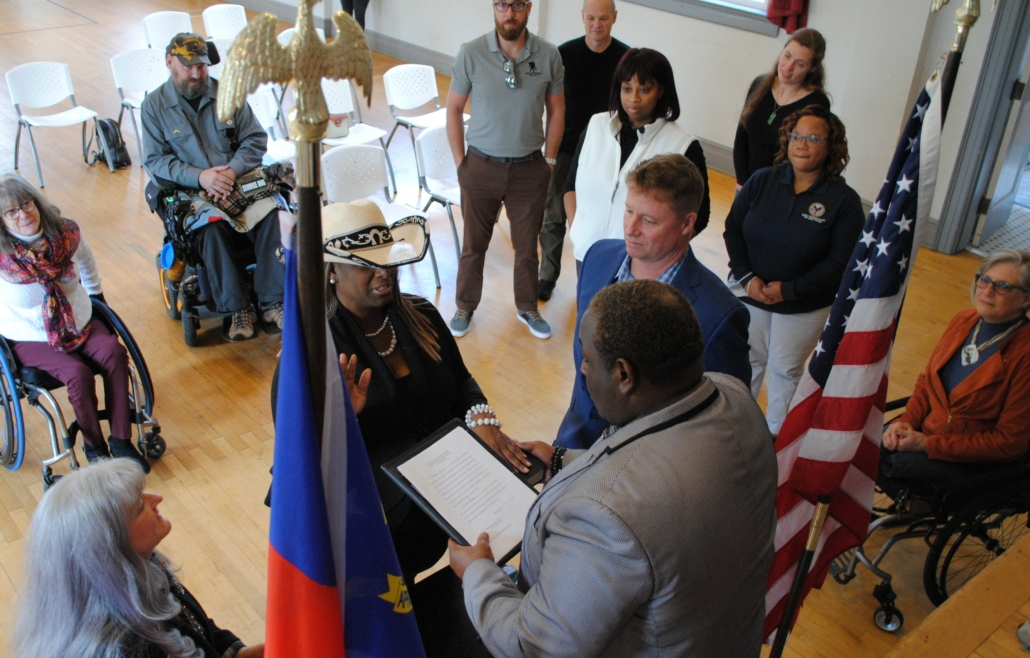 woman, Kanika Brown, in white cowboy had stands with right hand raised and left hand on bible. She is encircled by several people some standing and some in wheelchairs. On either side of Ms. Brown are the United States and North Carolina flags.